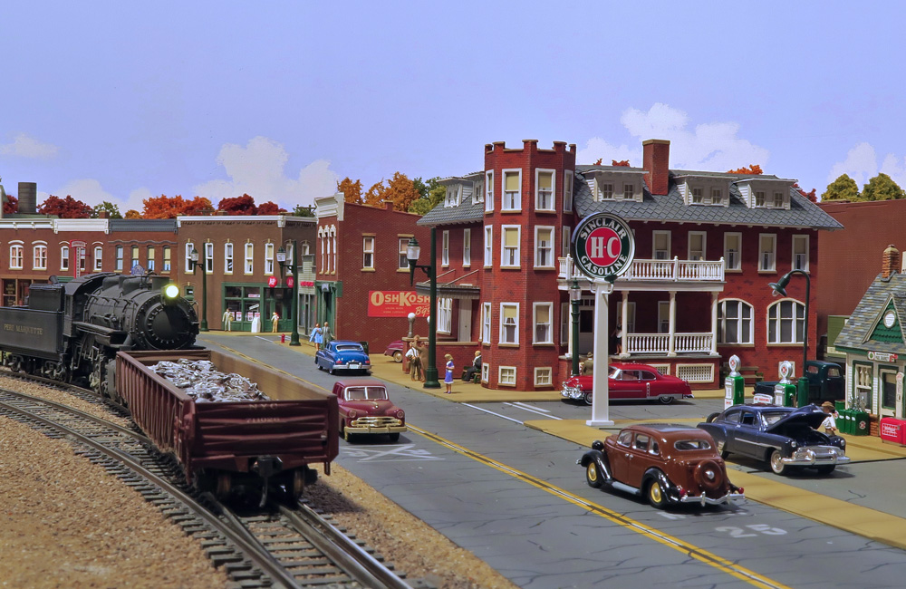 A steam engine switches a gondola of baled scrap metal next to an old-fashioned downtown scene