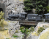 Dusty, black steam locomotive and tender haul passenger train over bridge into a tunnel.