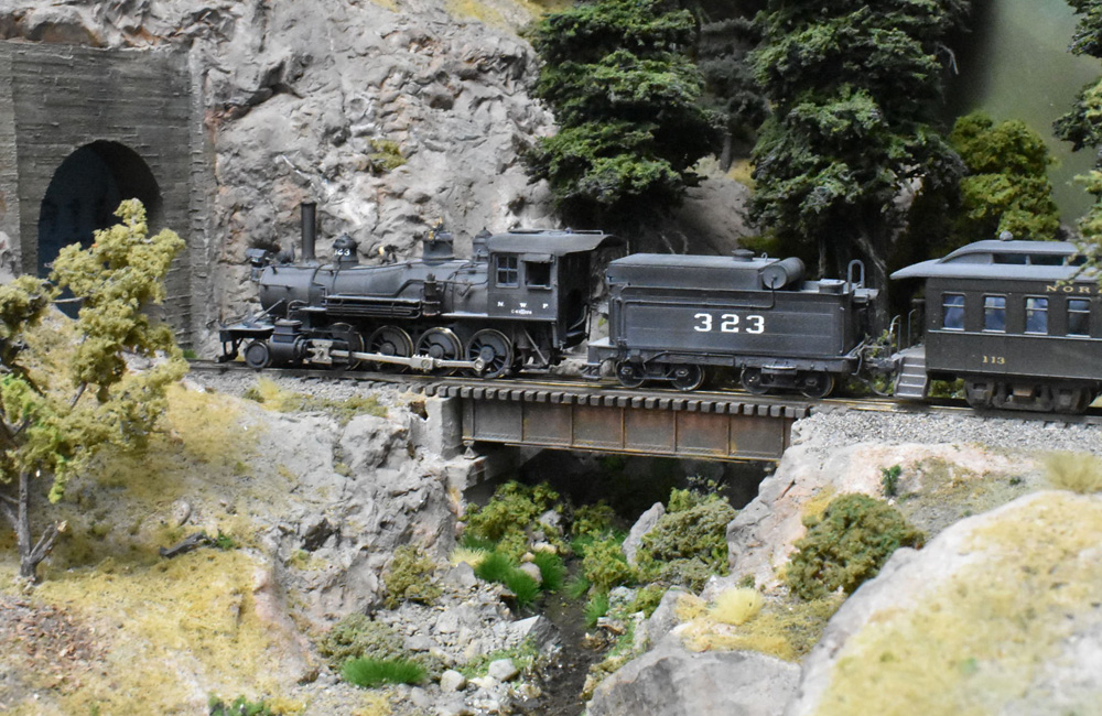 Dusty, black steam locomotive and tender haul passenger train over bridge into a tunnel.