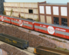 Three scarlet-and-gray 86-foot boxcars roll past a row of factories and a scrapyard