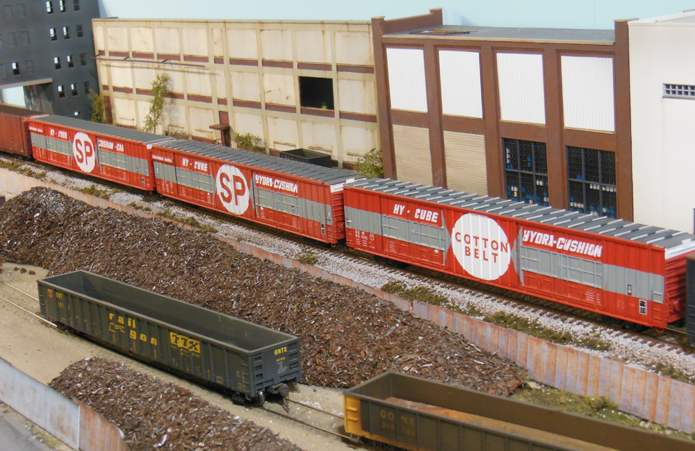 Three scarlet-and-gray 86-foot boxcars roll past a row of factories and a scrapyard