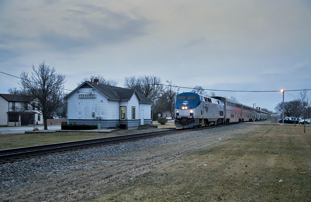 Passenger train passes through small town in dim pre-dawn light.