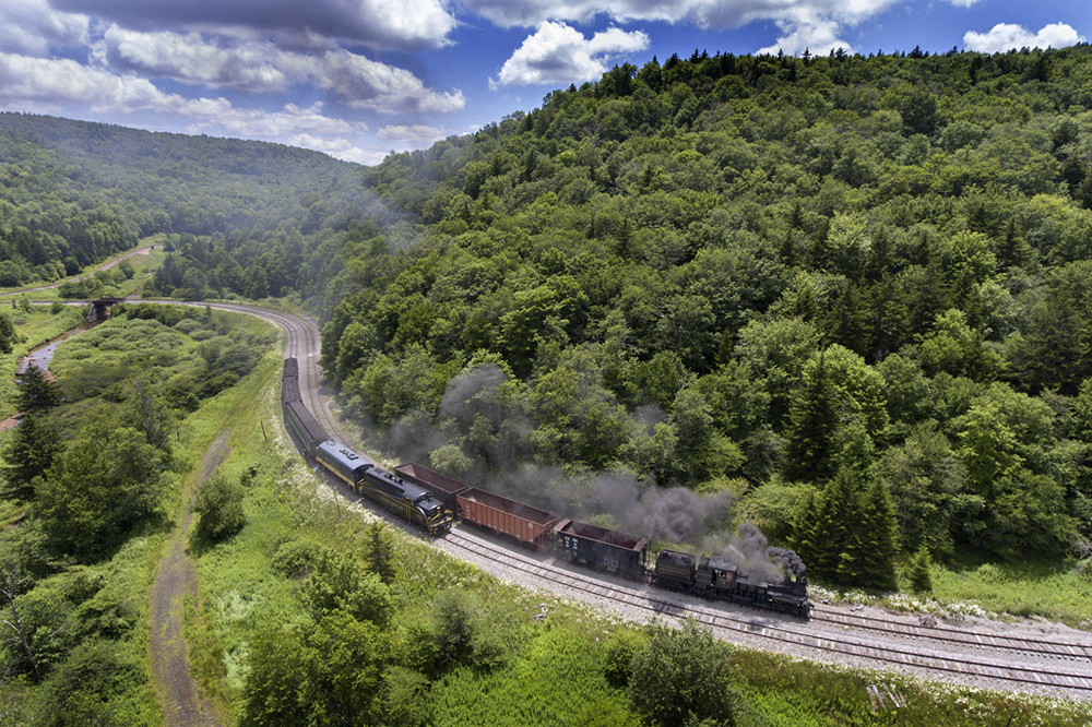 Silence on railfan photo lines: A high-angle image of a steam and diesel locomotive in a tree covered mountain landscape.