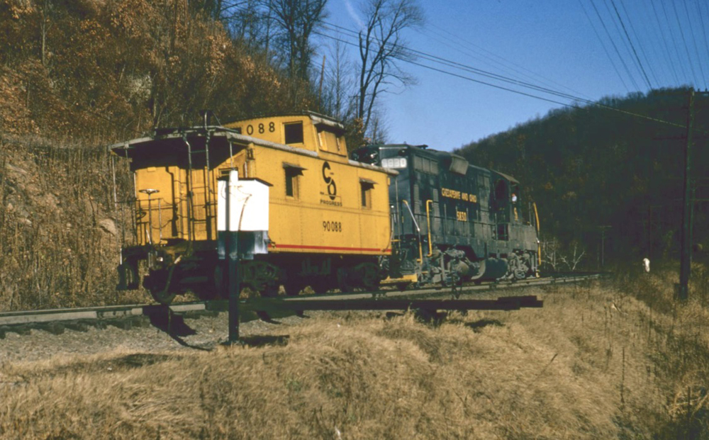 Locomotive and yellow caboose pass white phone box on pole