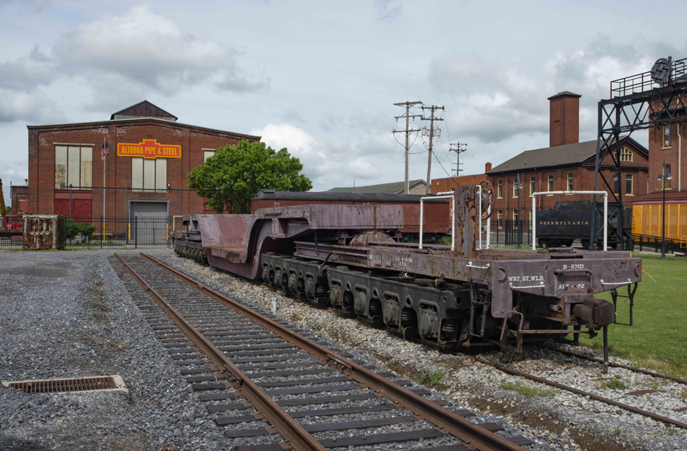 Depressed-center flat car with many wheels outside brick building