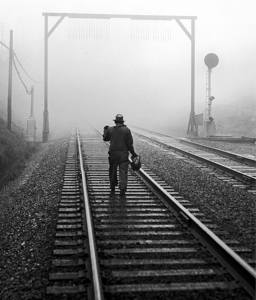 Black-and-white image of man walking down railroad tracks in fog
