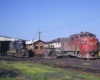 Red streamlined diesel locomotive with two black and white and one blue and yellow diesel locomotives in front of enginehouse