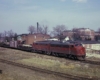 Maroon and red diesel locomotive with freight train in front of brick buildings