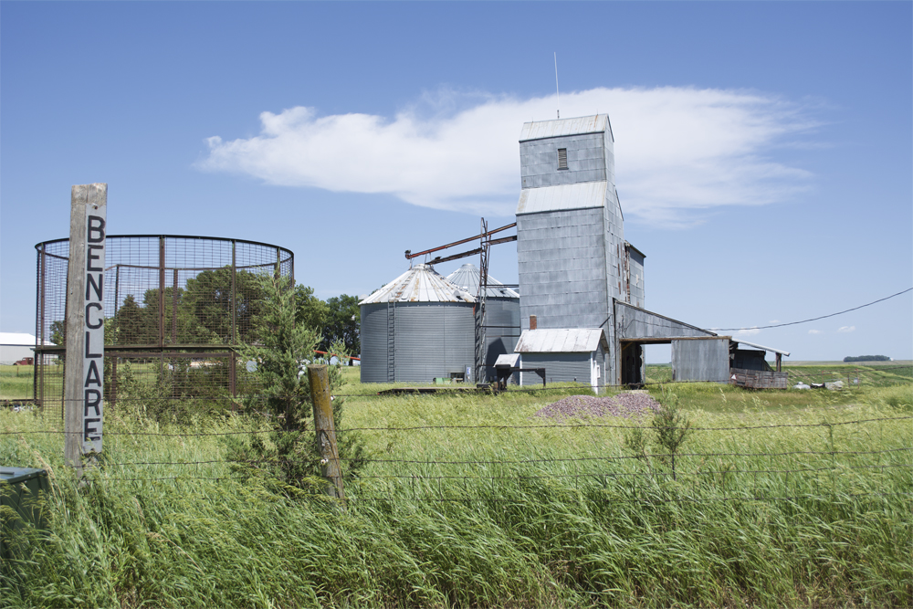 Photo with grain elevator, grain bins, corn crib, and sign on post