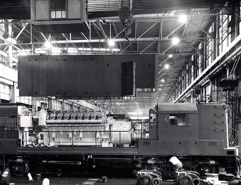Black and white image of a shell above a disassembled locomotive in a large workshop-warehouse.