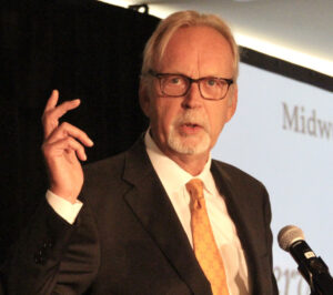 Man in dark suit and yellow tie speaking at microphone