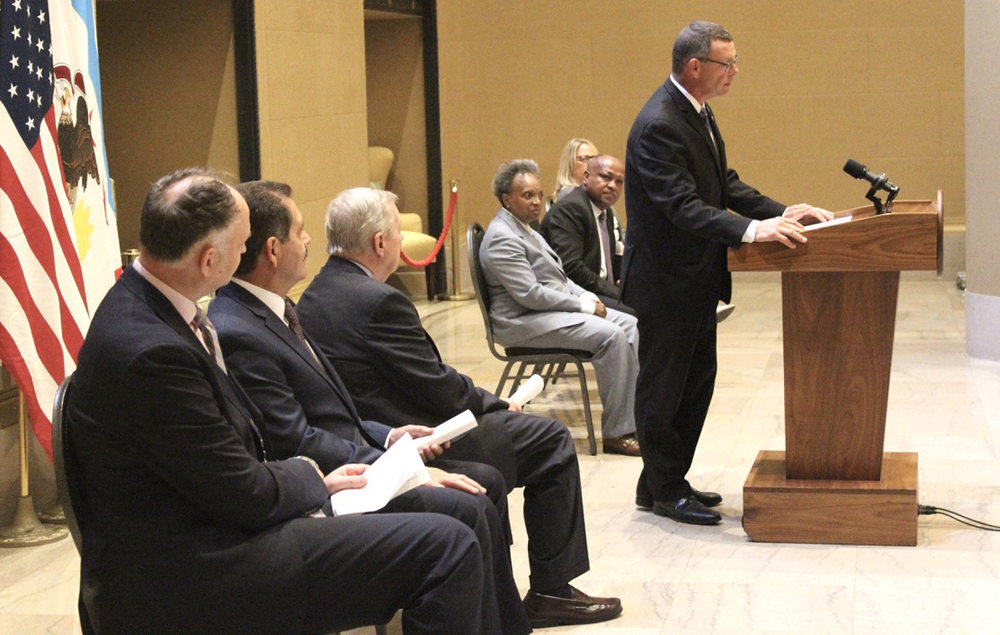 Man speaks at podium as other officials watch and listen from seats