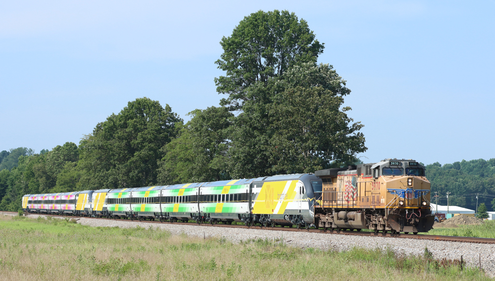 Green and pink passenger cars with yellow streamlined locomotives are pulled by a Union Pacific freight locomotive