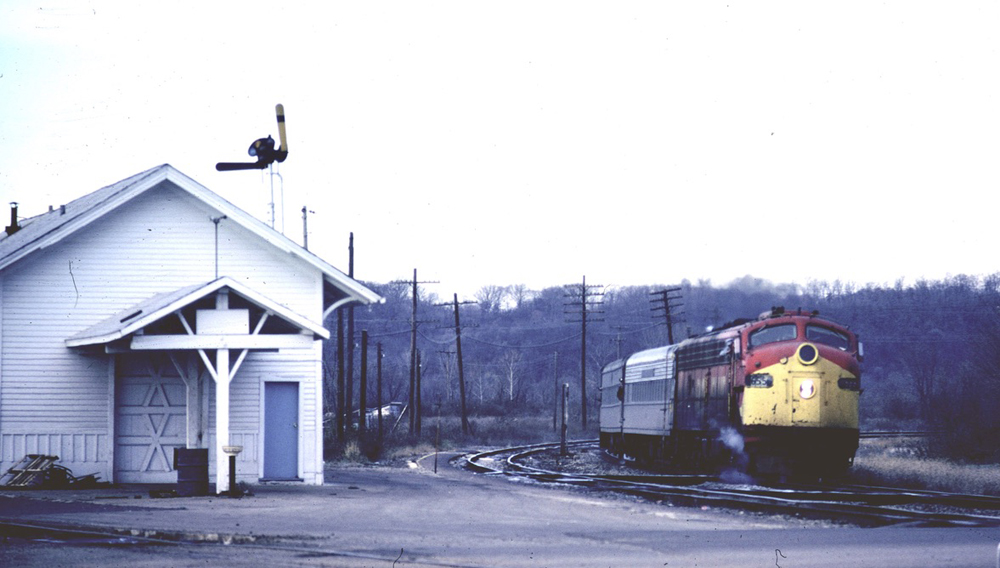 Short passenger train with red and yellow E-unit diesel passes small station