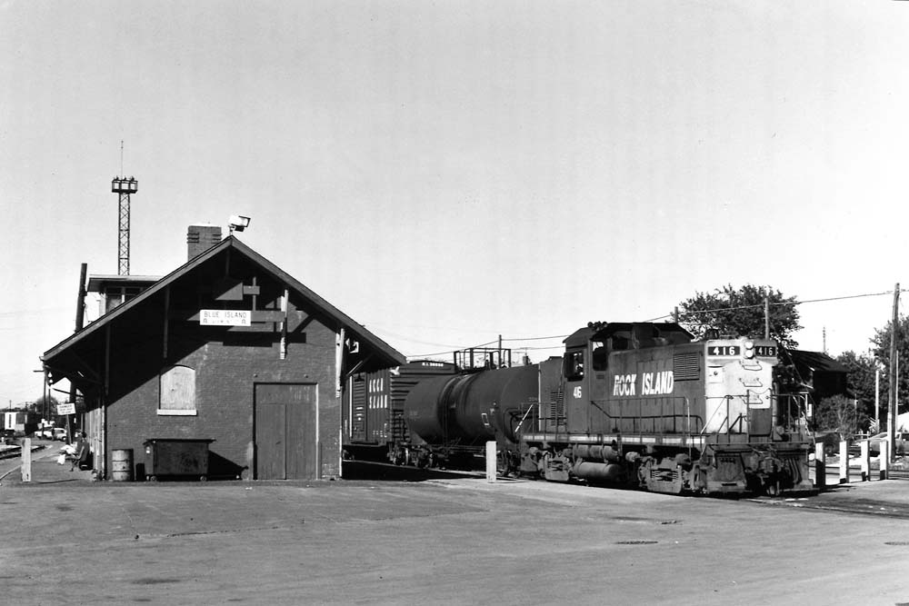 Center-cab diesel locomotive with freight cars passes brick station and open parking lot