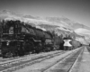 Diesel-powered freight train passes steam locomotive below mountains