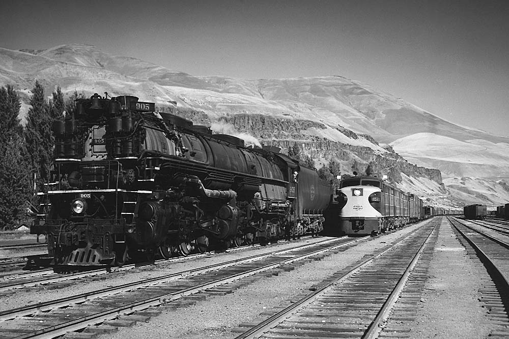 A diesel-powered freight train passes a steam locomotive under the mountains