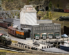 A steam locomotive pulls past a gray waterfront warehouse with a fishing boat docked nearby