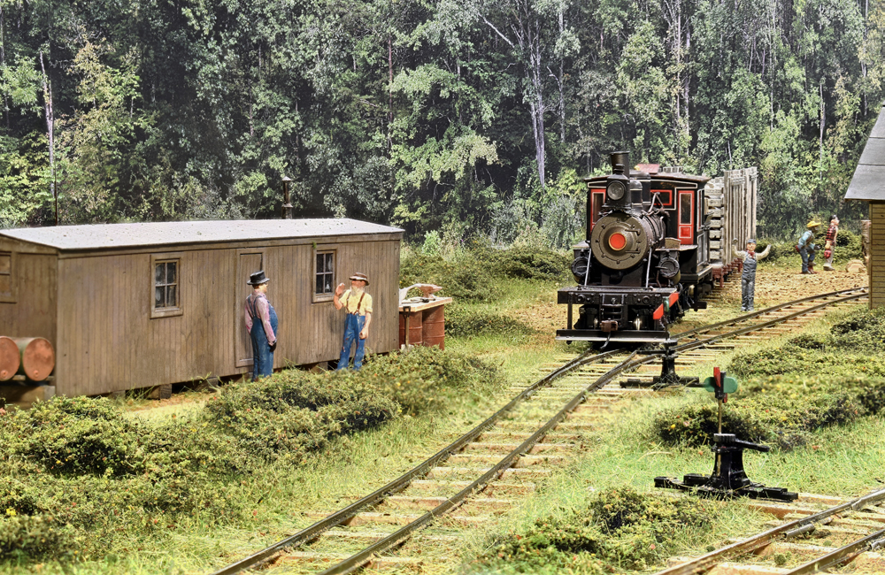 A narrow gauge geared steam locomotive heads toward the viewer through a logging camp with a pulpwood car in tow