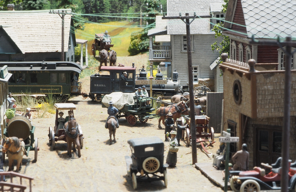 A small, black geared steam locomotive pulls a green wooden coach across a street bustling with pedestrians and horse-drawn wagons