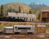 A silver Galloping Goose railcar rolls on the main line while behind it a black steam engine approaches a station