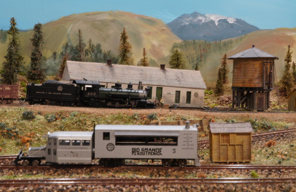 A silver Galloping Goose railcar rolls on the main line while behind it a black steam engine approaches a station