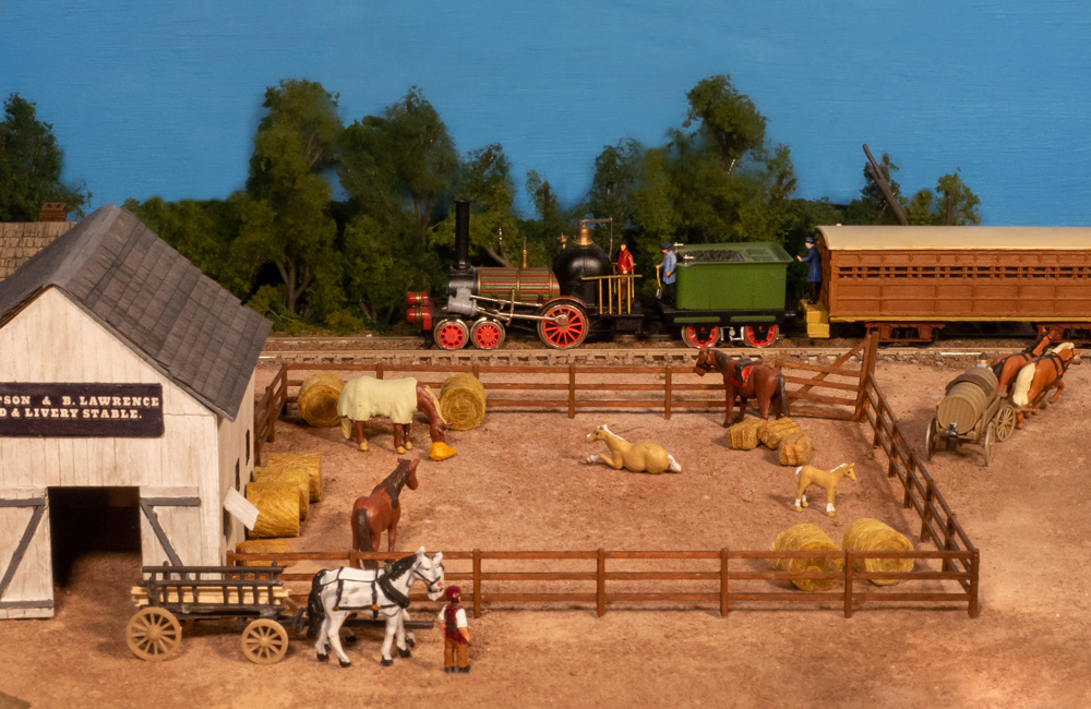 A small, old-fashioned 4-2-0 steam locomotive with a green tender pulls a wooden coach past a livery yard with horses