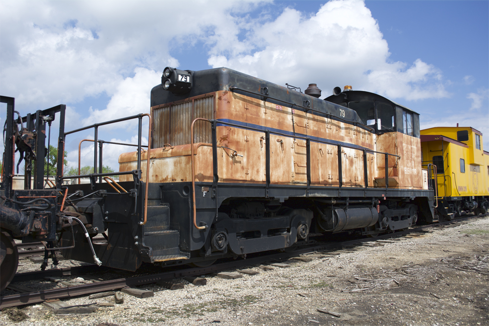 Photo of heavily weathered end-cab switcher with a yellow caboose in background.