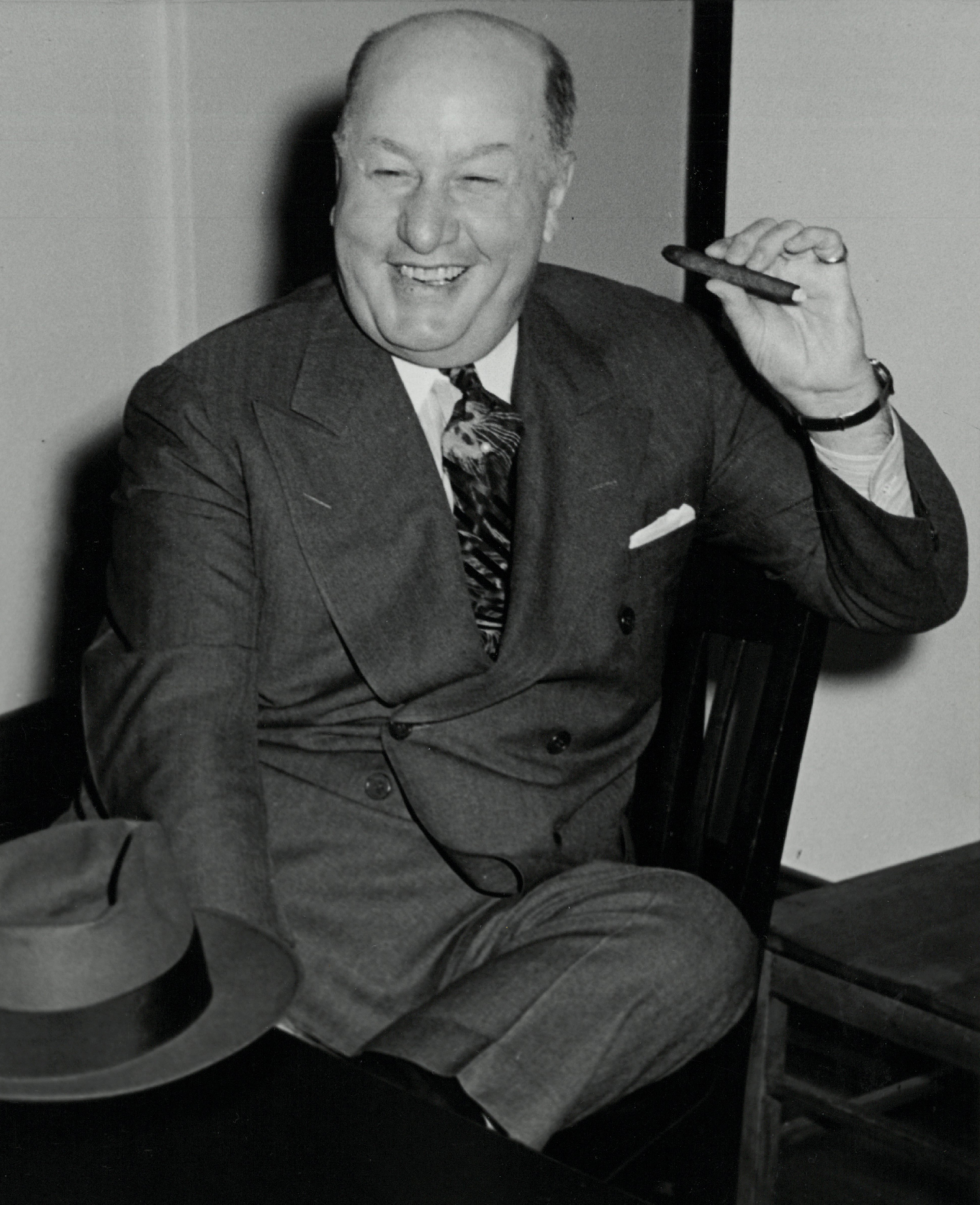 Bald man sitting on a chair, smoking a cigar. He has a smiling, happy face.