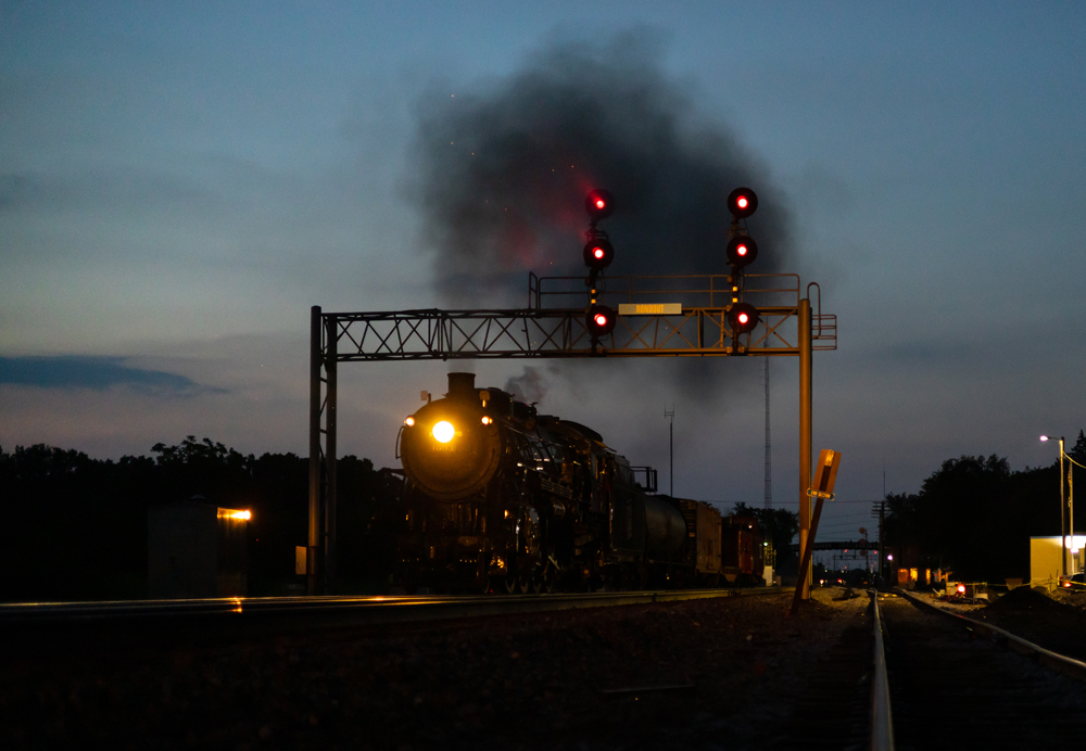 Steam locomotive passes under signal bridge at twilight