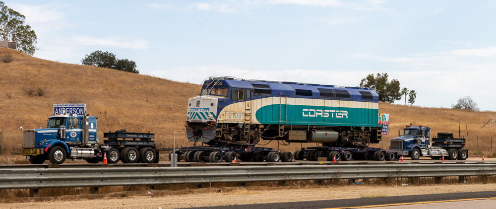 Blue, teal, and white locomotive on special wheeled trailer next to highway