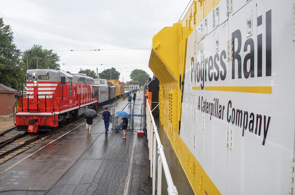 Red locomotive with train of cabooses next to yellow locomotive, as seen from on walkway of yellow locomotive