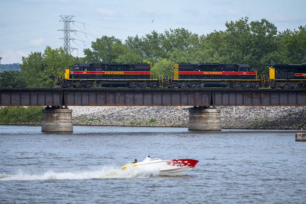 Black, red, and yellow locomotives cross bridge with speedboat in foreground
