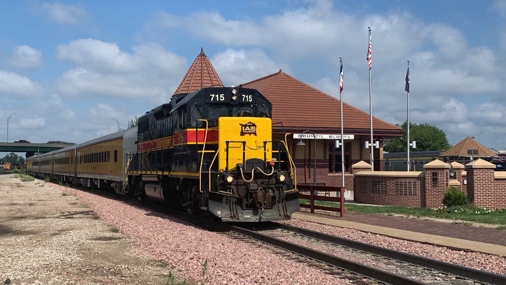 Black, red, and yellow locomotive with yellow passenger cars passing train station