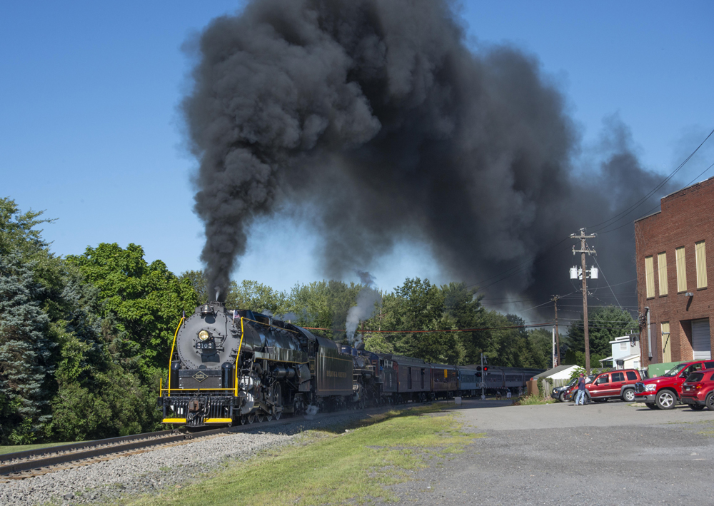 Two steam locomotives under large cloud of black smoke as they pass through a town