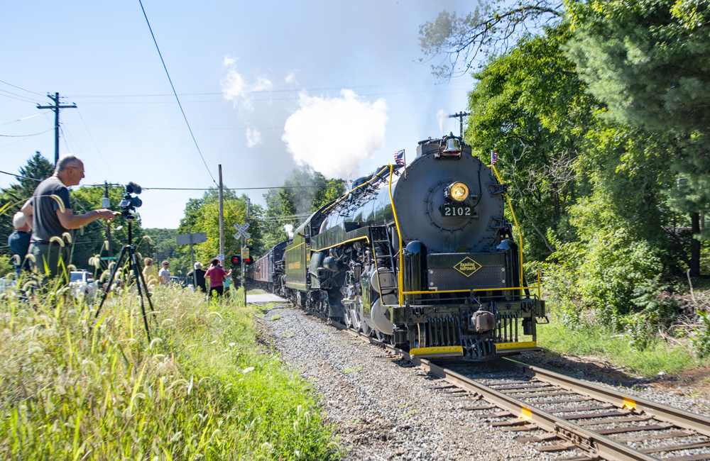 Train with steam locomotives passes spectators and man with video camera
