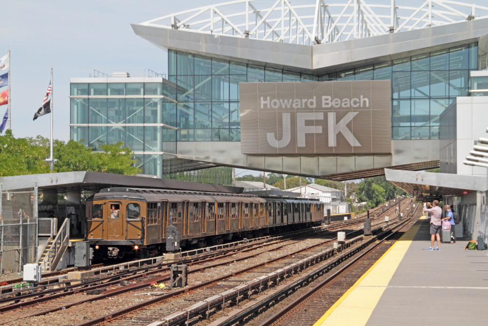 Vintage subway train passes under large glass station with "Howard Beach JFK" sign