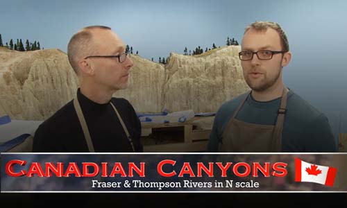 Two men in front of a mountainous model train layout with logo.