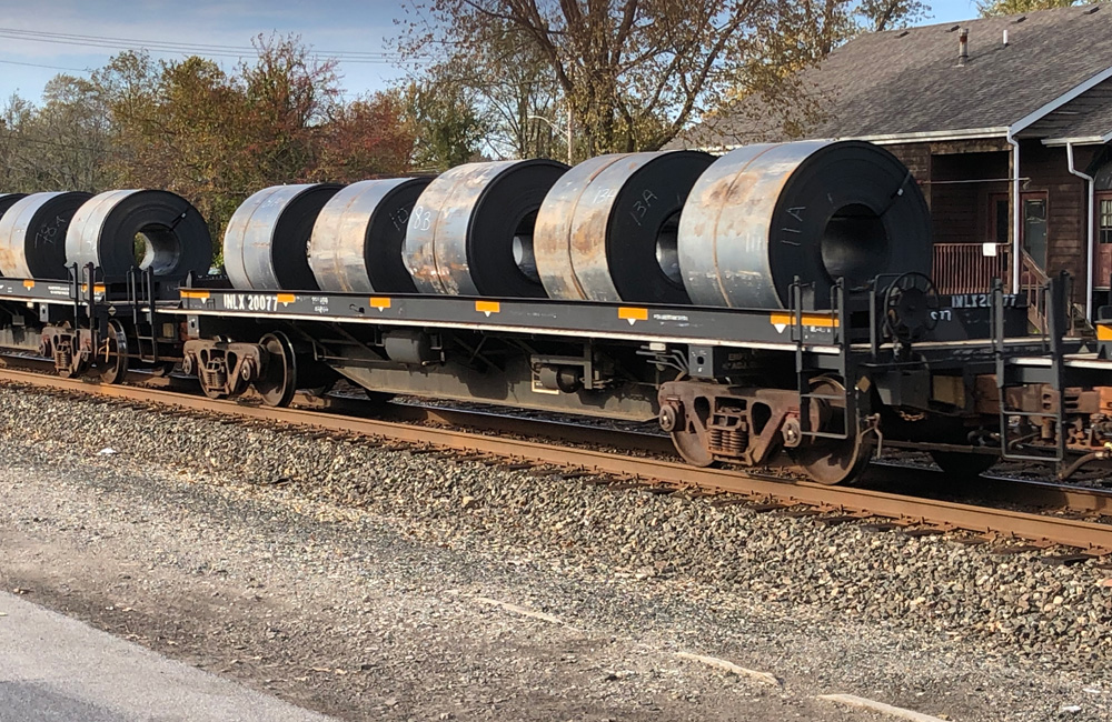 A black coil steel gondola car loaded with five steel coils rolls by a station in a freight train