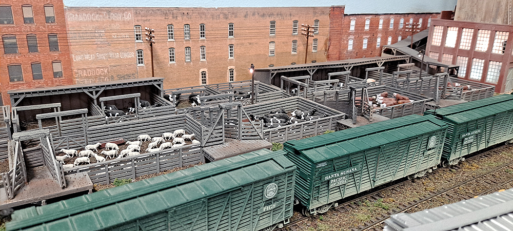Green Santa Susana Pacific stock cars are lined up in front of stockyard corrals with cows and sheep