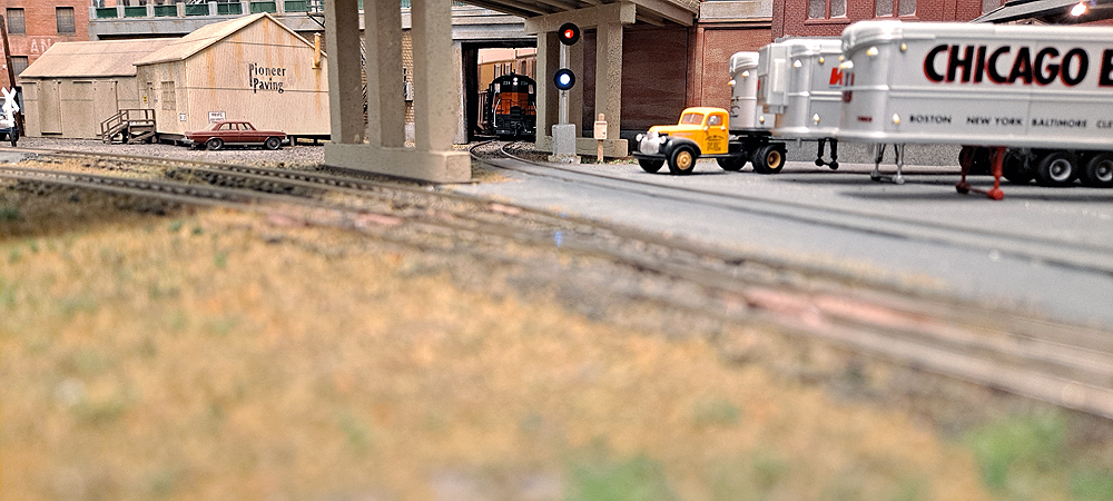 Tracks pass under highway overpass with trailer vans parked at right, red over green signal at center, with Milwaukee Road diesel emerging from tunnel through building at background