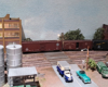 Silver vertical oil tanks in foreground, brick and concrete industrial building in background, black locomotive pushing three boxcars into spur