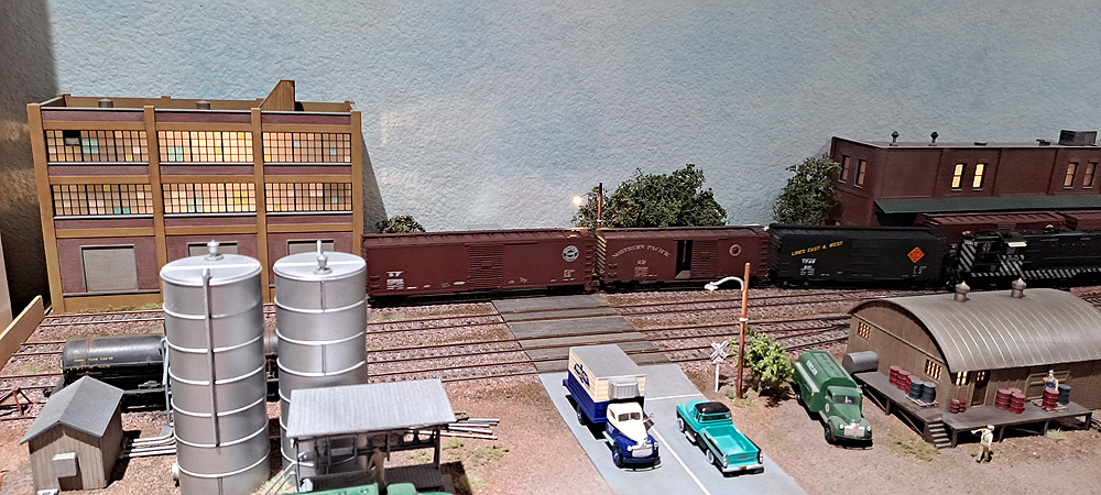 Silver vertical oil tanks in foreground, brick and concrete industrial building in background, black locomotive pushing three boxcars into spur