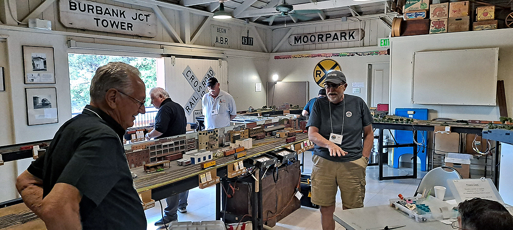 Wood frame railroad freight house, sliding freight door open to outside in background, modular train layout set up in midground surrounded by people looking at layout and chatting