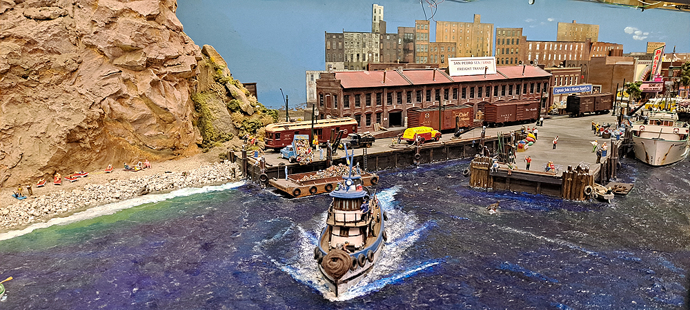 A tugboat leaves a dock across blue acrylic water with a rock bluff to the left and a brick warehouse on the rail-served wharf behind. Another wharf reaches out into the water at an angle