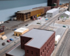 Two sets of parallel double track run through scene with brick building between the pairs. Road crosses tracks at 90-degree angle in midground, background, from left, has lumber distributor, large warehouse complex on far side of street. Vertical silver tanks, sawtooth-roofed building in background