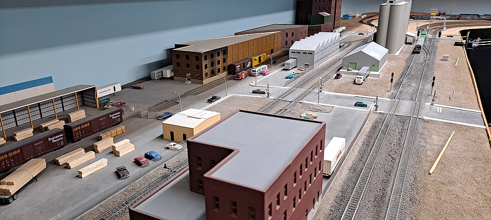 Two sets of parallel double track run through scene with brick building between the pairs. Road crosses tracks at 90-degree angle in midground, background, from left, has lumber distributor, large warehouse complex on far side of street. Vertical silver tanks, sawtooth-roofed building in background