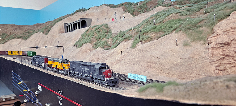 Three six-axle diesels in Southern Pacific, Union Pacific, and SP paint sit on siding in foreground as mountain rises in background. Rock shed near top of steep incline with more mountain above