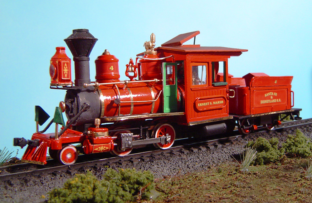 A scale model of a diminutive, shiny red steam locomotive with a four-wheel tender sits on a landscaped track