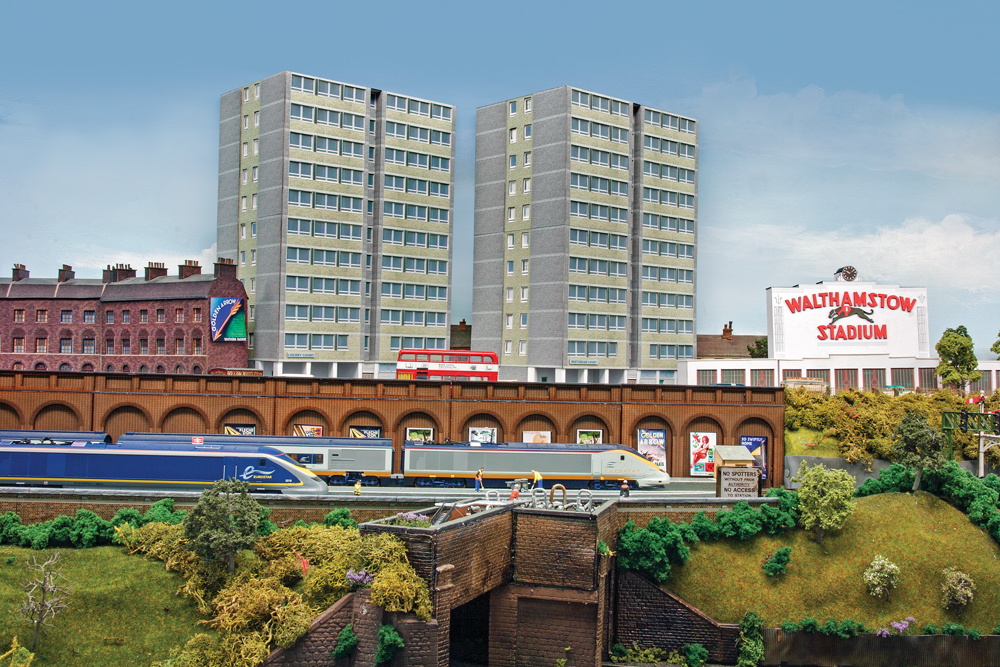 Two modern streamlined trains sit in front of older flat buildings.